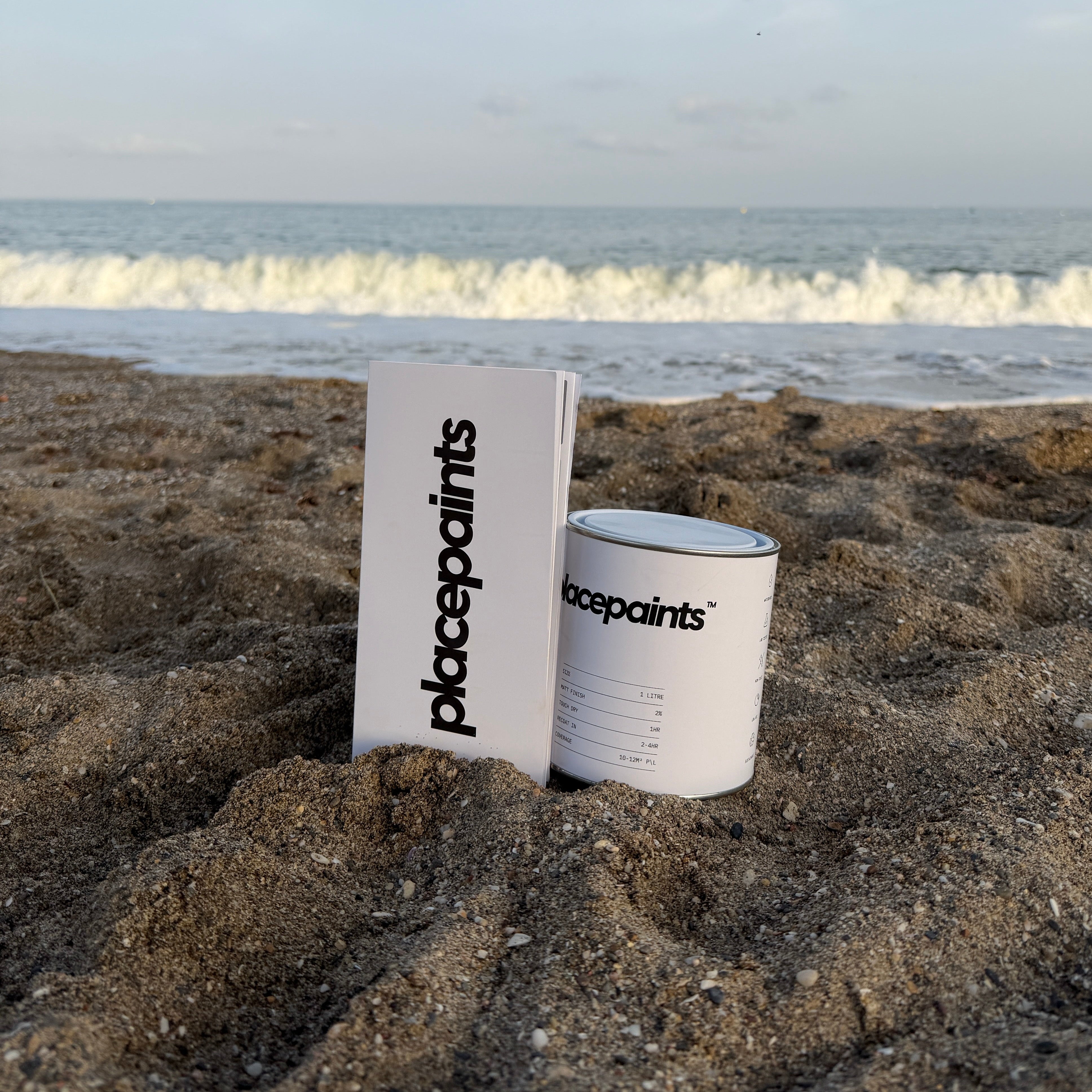 A white can of placepaints and a matching white box standing on a sandy beach with ocean waves and a blue sky in the background.